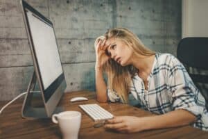 Junge Frau sitzt erschöpft am Schreibtisch vor einem Computer, stützt den Kopf in die Hand – Symbolbild für Überlastung, Stress und drohenden Burnout