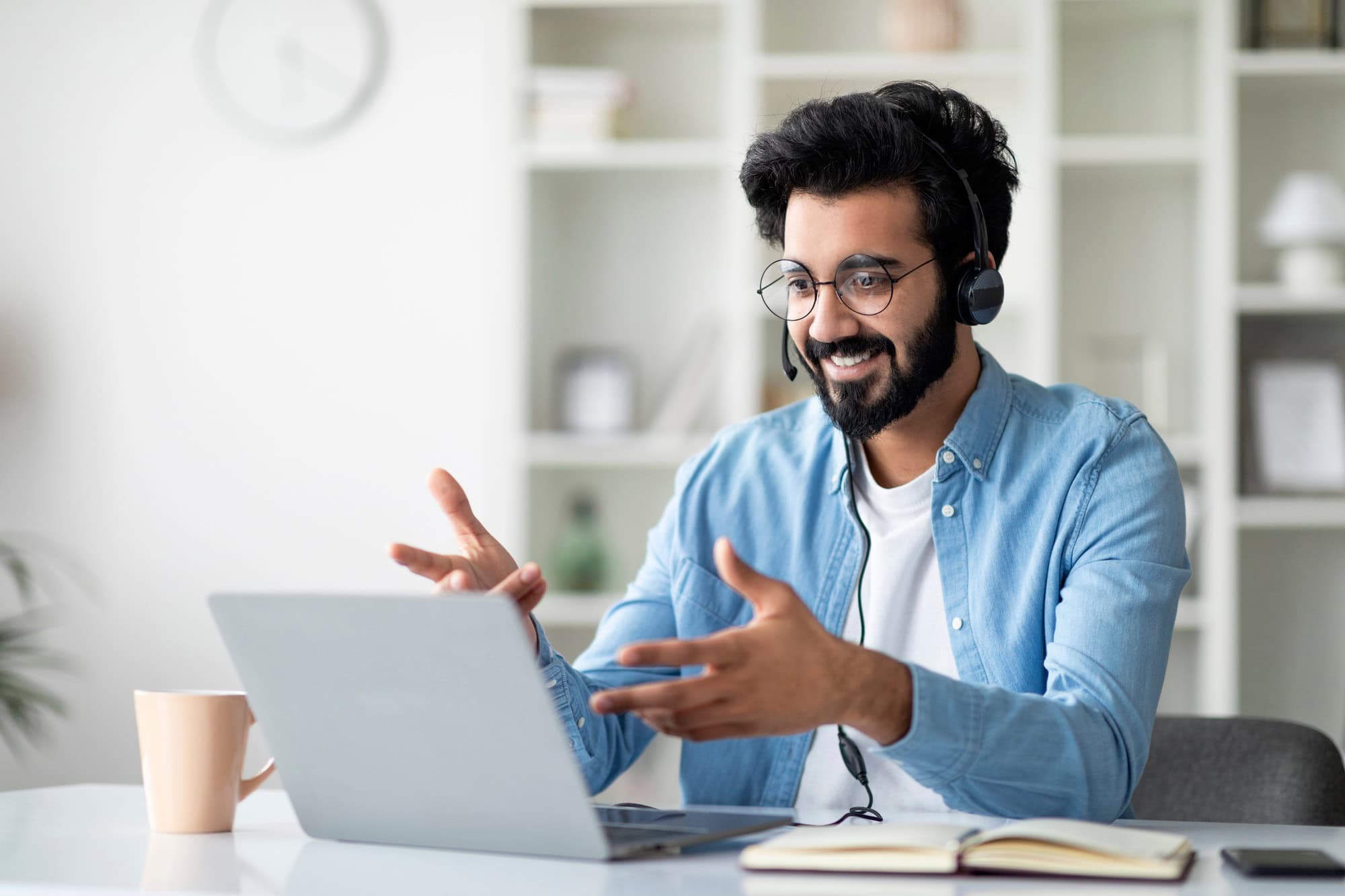 Mann beim Online-Coaching: Er sitzt mit Headset am Laptop, lächelt und gestikuliert in einem Gespräch, daneben ein Notizbuch