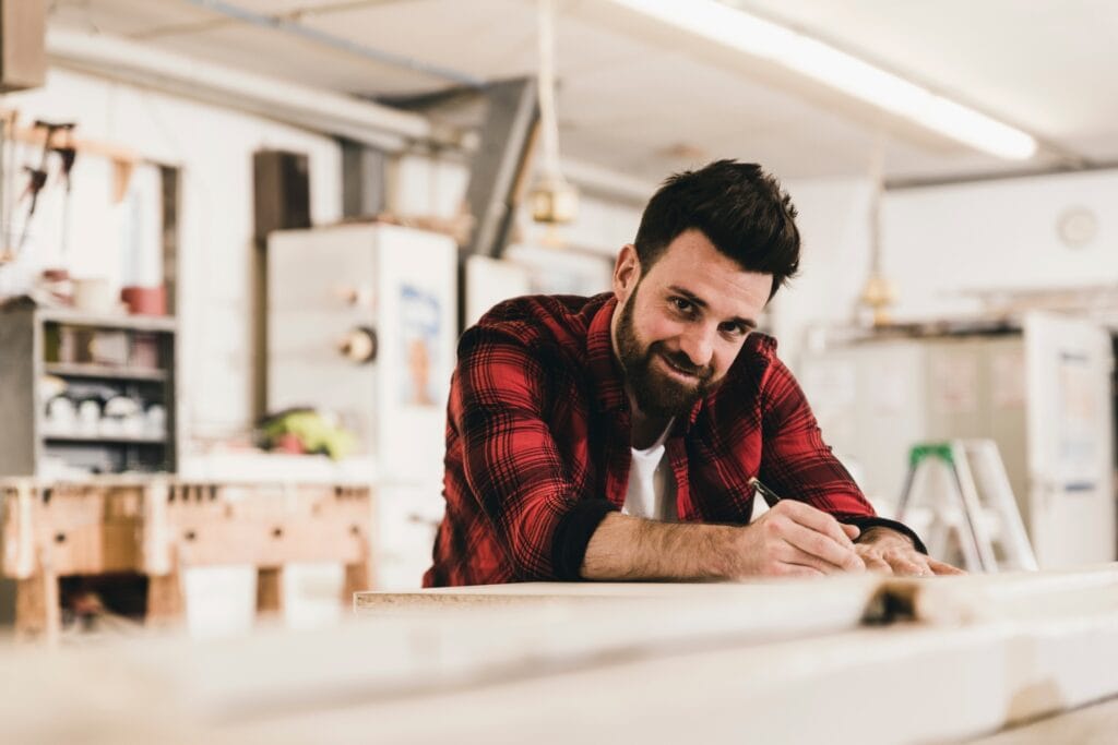 Handwerker in einer Werkstatt lehnt sich lächelnd auf die Werkbank – Sinnbild für gesunde, motivierte Mitarbeitende als Schlüssel für Unternehmenserfolg